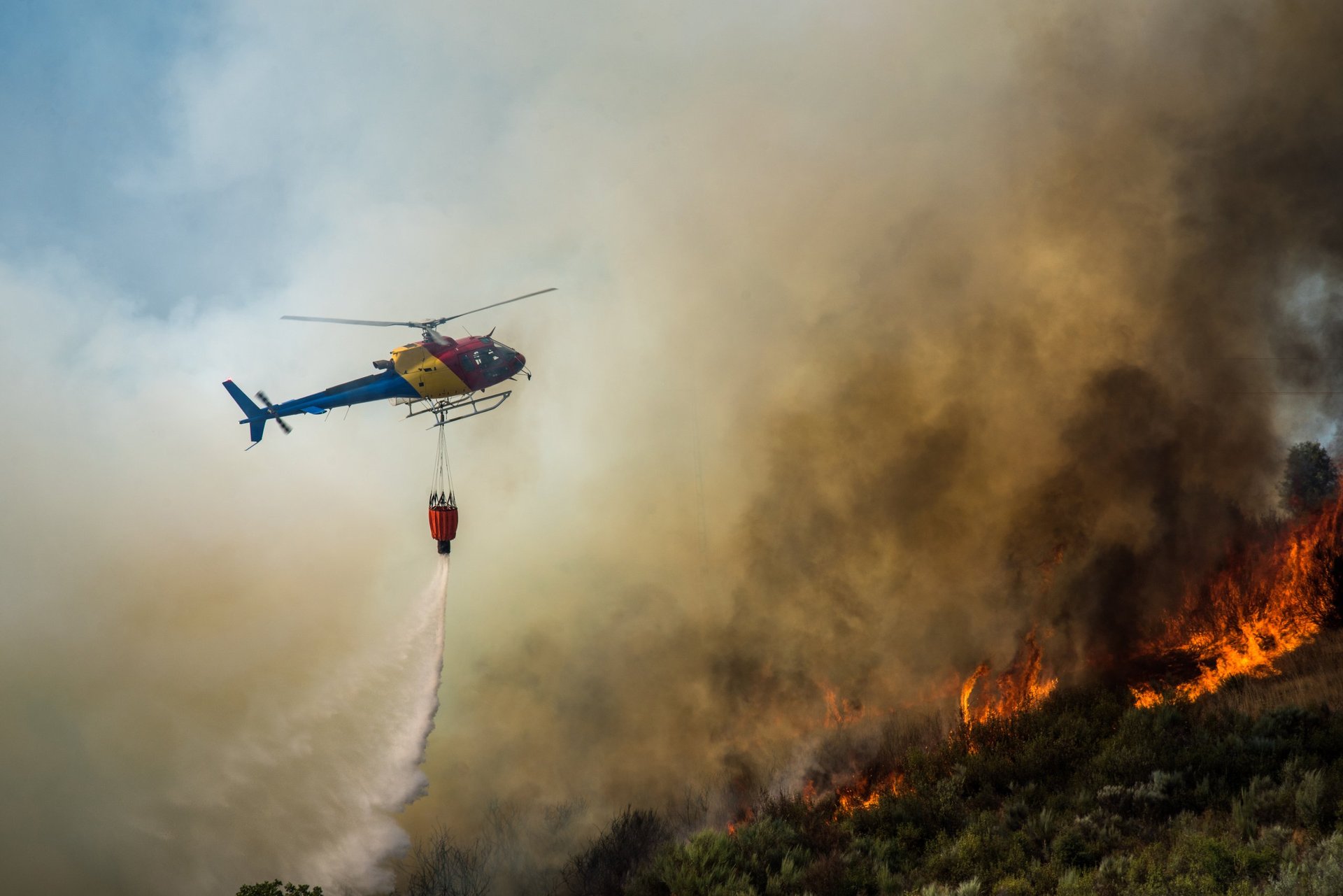 Helicopter dropping water above a forest fire in a mountainous region with thick smoke rising