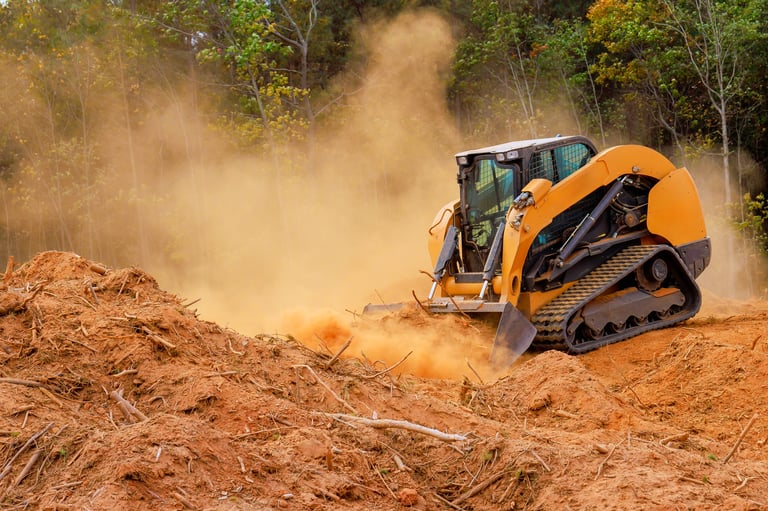 Skid steer works to clear plot of land in forest, kicking up dust moving soil