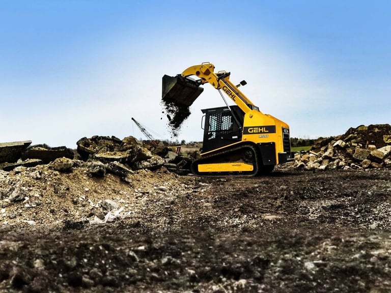 Yellow and black Gehl compact track loader operating on a construction site with dirt and debris piles under a blue sky