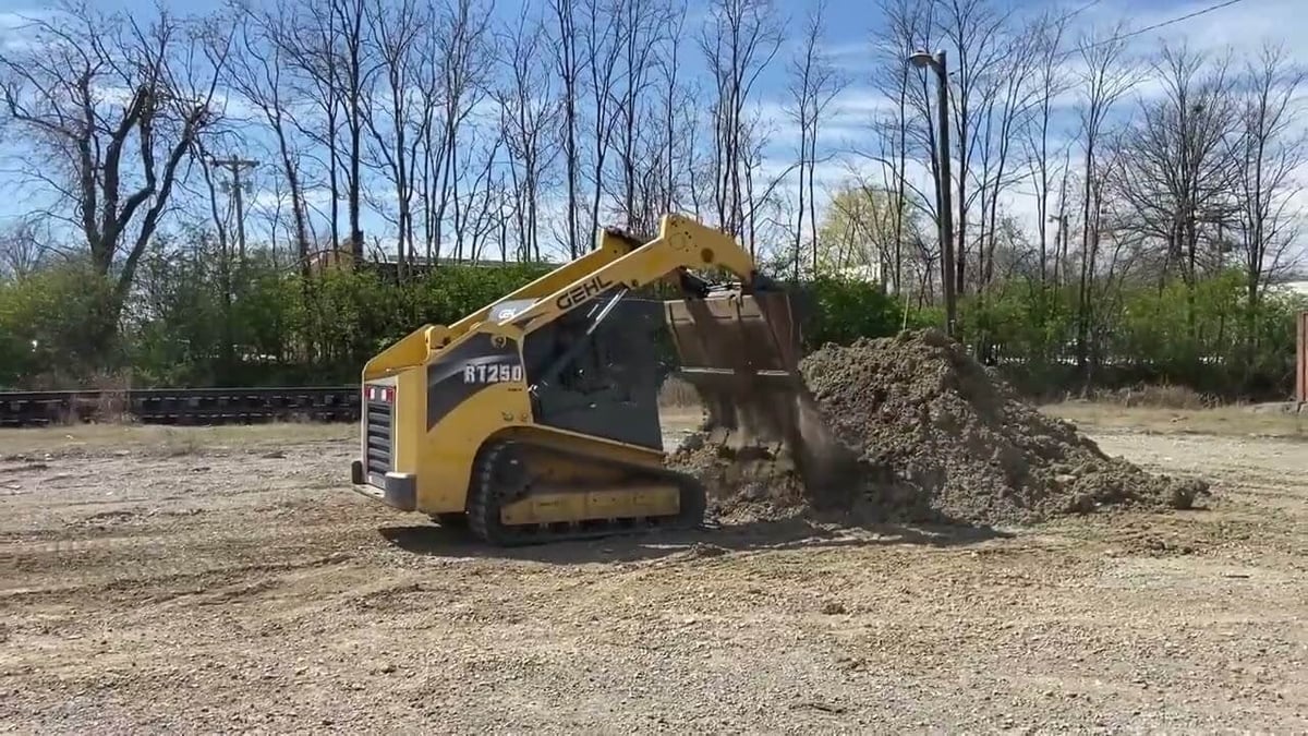 Yellow skid steer loader dumping gravel onto ground with bare trees in background