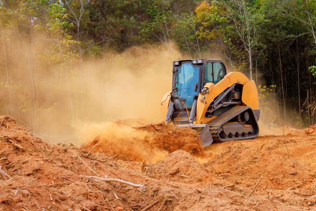 Orange and black tracked skid steer loader operating in a dusty clearing, moving earth and debris with trees in the background