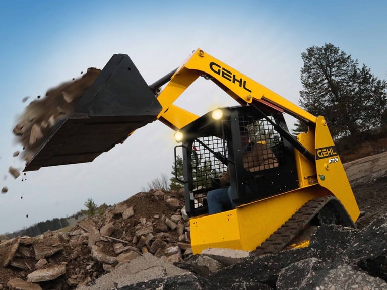 Yellow GEHL skid steer loader with raised bucket dumping dirt and rocks at a construction site