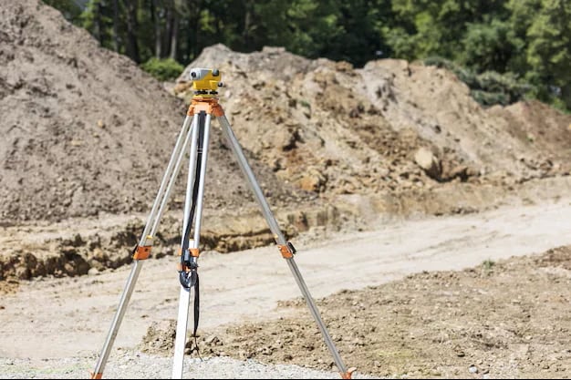Surveying equipment tripod with yellow optical level mounted on construction site with excavated earth piles in background