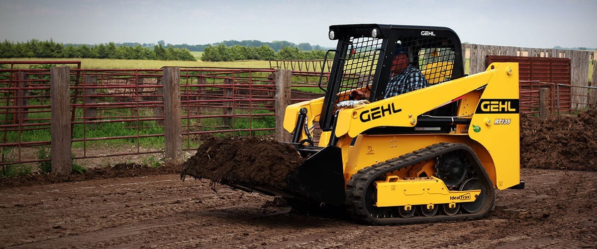 Yellow Gehl skid steer loader moving dirt near wooden ranch fencing in a farm field
